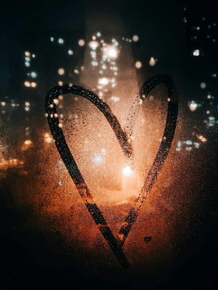 Heart shape drawn in condensation on a window, with a dark city scene behind it.